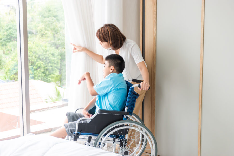 asian woman taking care disabled child on wheelchair in bedroom. both looking at something beside of window.